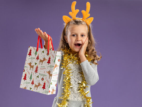 穿兴奋的小女孩戴着圣诞发箍 脖子上戴着花环 伸出礼品袋 手放在脸上 隔离在蓝色的墙上放箍花环