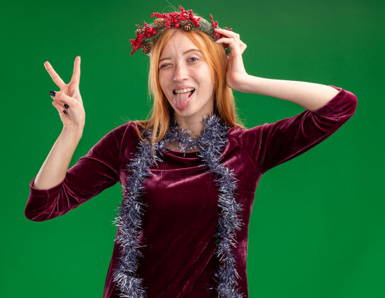 圣诞节快乐的眨眼年轻漂亮的女孩穿着红色的衣服 脖子上戴着花环和花环 在绿色的墙上显示出舌头和和平的姿态展示脖子年轻