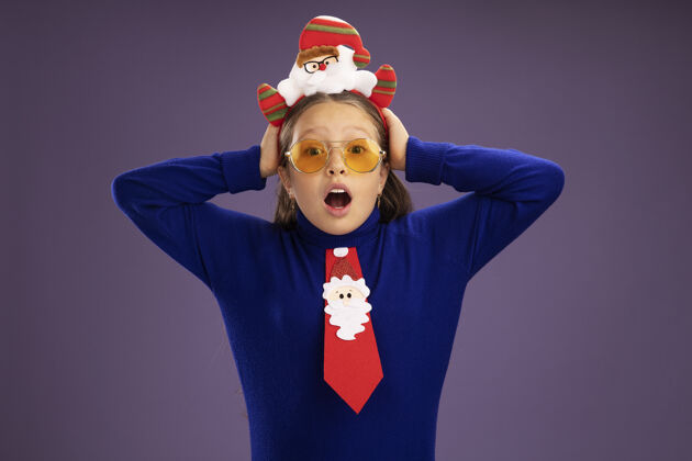 站着小女孩穿着蓝色高领毛衣 打着红色领带 头上戴着有趣的圣诞戒指 手放在头上 既担心又惊讶蓝色高领毛衣紫色