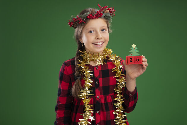花环快乐的小女孩戴着圣诞花环 穿着格子裙 脖子上挂着金属丝 手里拿着玩具立方体 脸上带着微笑 还有圣诞节的日期围着衣服脸