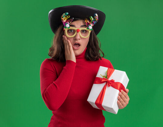 帽子穿着红毛衣 戴着滑稽眼镜 戴着黑帽子 拿着礼物的年轻女孩站在绿色的墙上 惊喜而快乐毛衣圣诞节惊喜