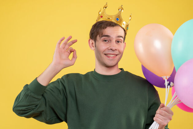 成人正面图帅气的年轻人戴着皇冠拿着气球在黄色上做奥基标志英俊气球气球
