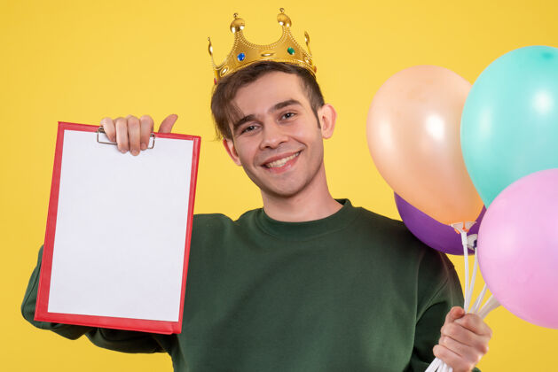 脸正面图帅气的年轻人戴着皇冠拿着气球和黄色的剪贴板英俊的年轻人剪贴板肖像