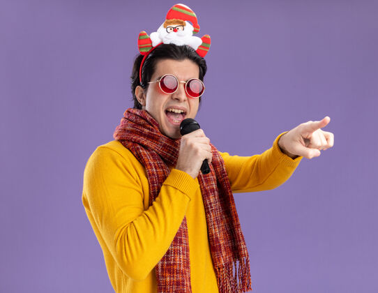 圣诞节一个快乐的年轻人 脖子上围着一条暖和的围巾 穿着黄色高领毛衣 头上戴着一副滑稽的眼镜 拿着麦克风唱歌 用食指指着紫色墙上站着的东西指向站着男人