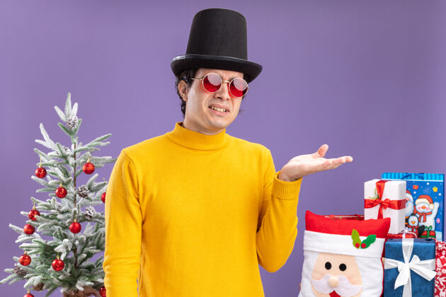 穿着一个穿着黄色高领毛衣 戴着眼镜 戴着黑色帽子 胳膊伸出来的年轻人正困惑地站在一棵圣诞树旁边 在紫色的墙上送礼物是年轻高领毛衣