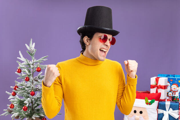 圣诞节身穿黄色高领毛衣 戴着黑色帽子的戴眼镜的年轻人站在圣诞树旁 兴奋地握紧拳头 在紫色的墙上送礼物兴奋男人年轻