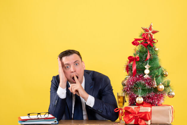 惊讶的男人前视图惊讶的人坐在圣诞树附近的桌子上 黄色的礼物办公室树商人