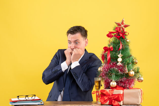 礼物困惑的男人坐在圣诞树旁的桌子前 呈现黄色坐着黄色桌子