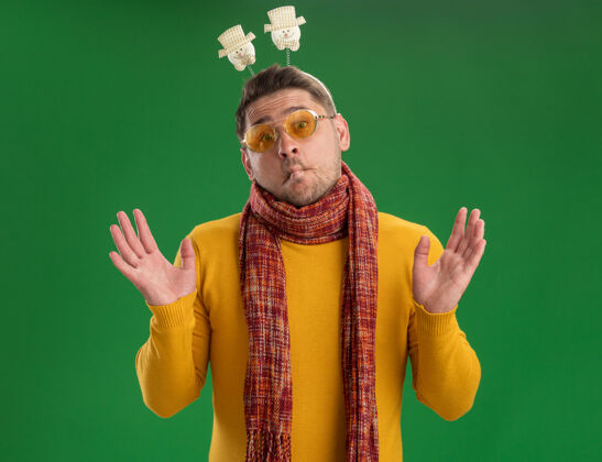 圣诞节一个穿着黄色高领毛衣戴着眼镜的年轻人 头上戴着一条暖和的围巾 站在绿色的墙上边缘年轻头
