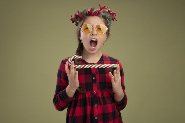 圣诞节快乐和兴奋的小女孩戴着圣诞花环在格子裙拿着糖果手杖看着相机尖叫站在绿色的背景衣服糖果尖叫