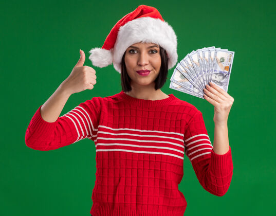 帽子高兴的年轻女孩戴着圣诞帽拿着钱 在绿色的墙上孤立地竖起大拇指展示年轻穿着
