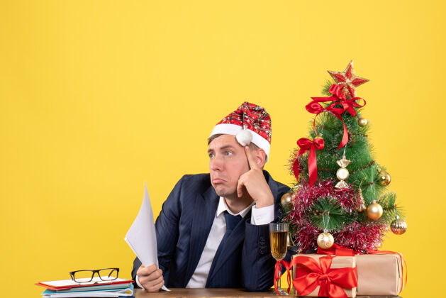 西装前视图：体贴的男人坐在圣诞树旁的桌子旁 黄色的礼物礼物人新郎