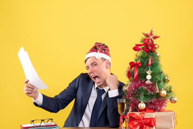 成人前视图兴高采烈的男子眨着眼睛坐在圣诞树旁边的桌子上 黄色的礼物套装圣诞节坐着