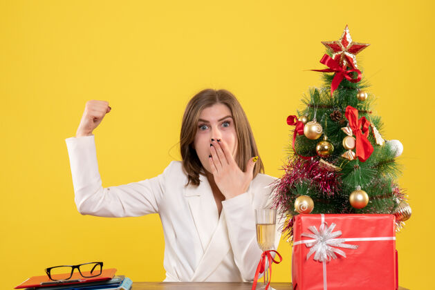 背景正面图：女医生坐在黄色背景的桌子前 放着圣诞树和礼品盒快乐肖像坐