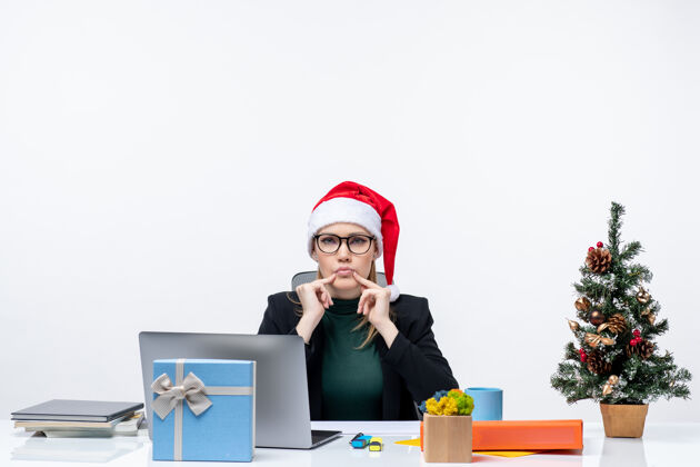 商务女士带着圣诞老人帽子的女商人坐在桌子旁 桌上放着圣诞树和礼物笔记本电脑电脑坐着