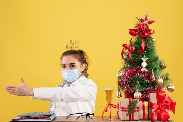 前面正面图女医生戴着黄色背景的无菌口罩坐在圣诞树和礼盒里面具圣诞节人
