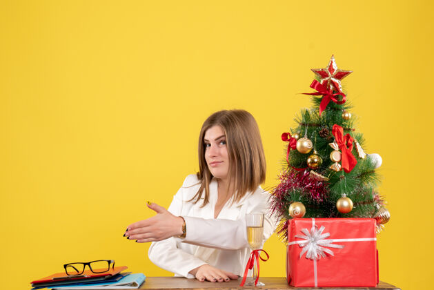 医院正面图：女医生坐在桌子前 拿着圣诞树和礼盒在黄色背景上握手手家成人