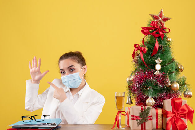 情感正面图女医生戴着防护面具坐在黄色背景上 戴着圣诞树和礼品盒医生病毒圣诞节
