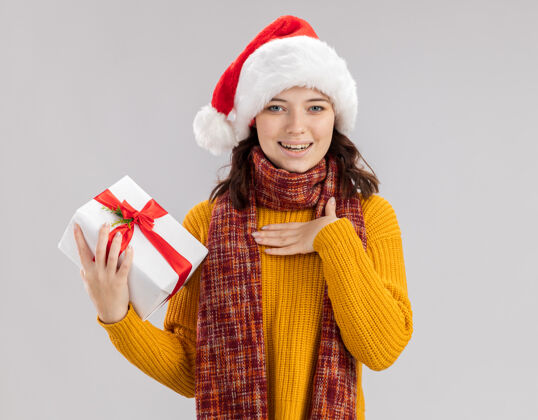 举行戴着圣诞帽 脖子上围着围巾的年轻斯拉夫女孩高兴地把手放在胸前 拿着圣诞礼盒 白色背景上有复制空间放礼物年轻人