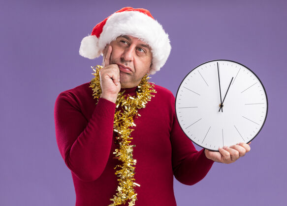 看中年男子戴着圣诞老人的帽子 脖子上戴着金属丝 拿着钟 站在紫色的背景上困惑地看着一边帽子金属片站着