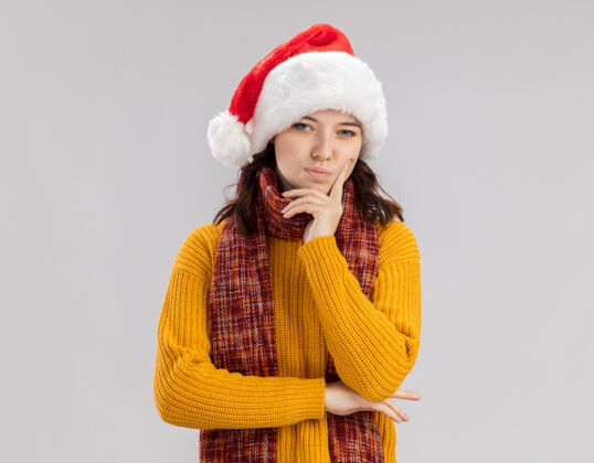 斯拉夫体贴的年轻斯拉夫女孩戴着圣诞帽 脖子上围着围巾 抱着下巴 看着隔离在白色背景上的相机 还有复印空间周围看起来背景