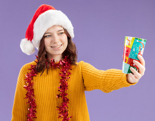 新的微笑着的年轻斯拉夫女孩 戴着圣诞帽 脖子上戴着花环 手里拿着纸杯 看着紫色背景上的复制空间脖子圣诞快乐圣诞老人