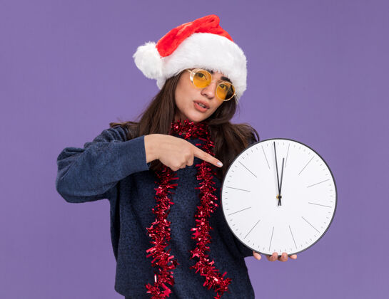 背景戴着太阳眼镜 戴着圣诞帽 脖子上戴着花环的自信的年轻白种人女孩拿着并指着紫色背景上的时钟 背景是复制空间时钟圣诞快乐年轻