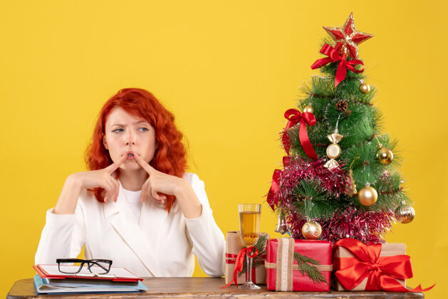圣诞节正面图：女医生坐在桌子后面 拿着圣诞礼物和黄色背景上的树思考人礼物思考