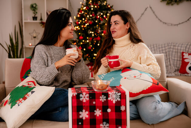 高兴高兴的年轻女孩们拿着杯子 看着彼此坐在扶手椅上 在家里享受圣诞节的时光漂亮扶手椅享受