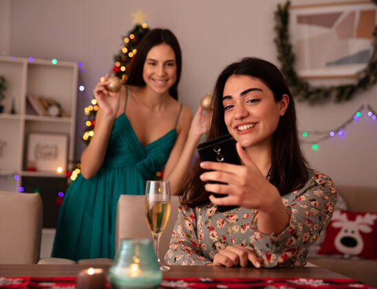 快乐笑容可掬的年轻女孩拿着玻璃球饰品站在朋友身后自拍 坐在餐桌旁 在家里享受圣诞节时光年轻漂亮家