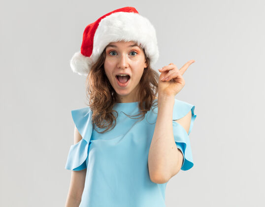 惊讶穿着蓝色上衣 戴着圣诞帽的年轻女子看起来很惊讶 聪明的脸上带着微笑 有一个新的好主意 伸出食指顶部有年轻