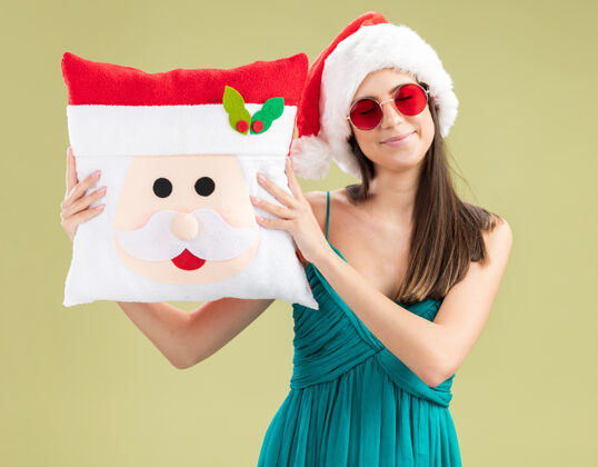 复制戴着太阳眼镜 戴着圣诞帽 抱着圣诞枕头的年轻白种女孩很高兴橄榄年圣诞快乐