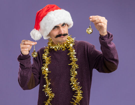 帽子小胡子男人戴着圣诞帽子 脖子上戴着金属丝 手里拿着圣诞球 站在紫色的背景下 带着怀疑的表情看着摄像机脖子表情站着
