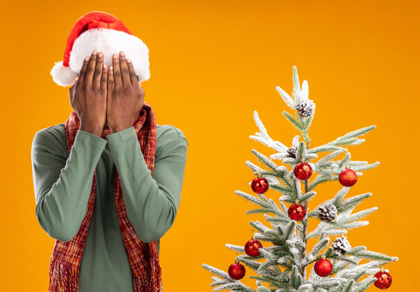 掩护戴着圣诞帽 围着围巾的非洲裔美国人站在橙色墙上的圣诞树旁 用手遮住脸围着圣诞老人围巾