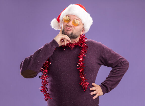 腰令人印象深刻的中年男子戴着圣诞老人帽 脖子上戴着金箔花环 戴着眼镜 一边看着一边 手放在腰上 下巴孤立在紫色的墙上下巴保持帽子