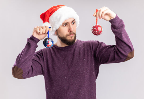 圣诞节一个穿着紫色毛衣 戴着圣诞帽 戴着滑稽眼镜 拿着圣诞球的年轻人站在白色的墙上困惑地看着他们 试图做出选择站着球男人