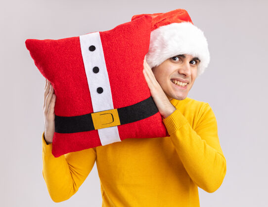 高领毛衣穿着黄色高领毛衣 戴圣诞帽的年轻人抱着圣诞枕头 站在白色的墙壁上欢快地笑着高兴地圣诞老人男人