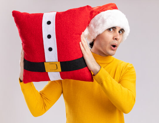 惊讶一个穿着黄色高领毛衣 戴着圣诞帽 抱着圣诞枕头的年轻人站在白色的墙上 惊讶又惊奇枕头高领毛衣惊讶