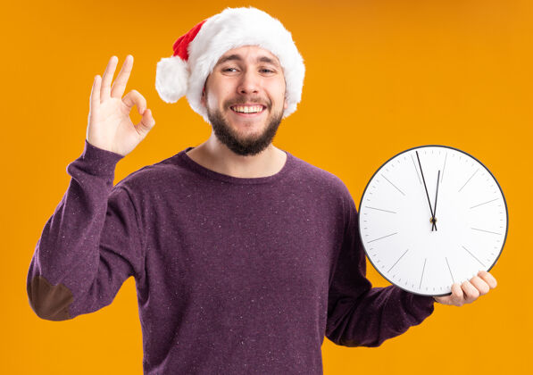 年轻穿着紫色毛衣 戴着圣诞帽的年轻人拿着挂钟 高兴地站在橙色的墙上 露出ok的微笑时钟展示毛衣