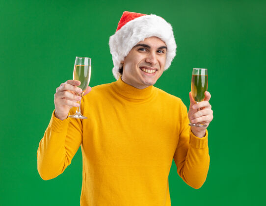 圣诞老人穿着黄色高领毛衣 戴着圣诞老人帽的快乐的年轻人举着两杯香槟 站在绿色的墙上欢快地笑着帽子兴高采烈年轻