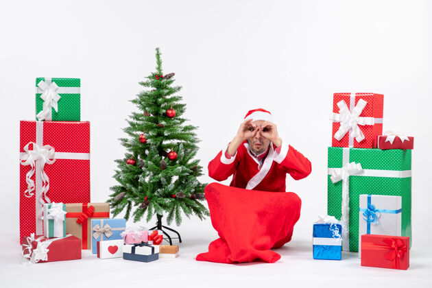 礼物年轻人庆祝圣诞节假期坐在地上做眼镜手势 靠近礼物和圣诞树装饰圣诞树礼物眼镜
