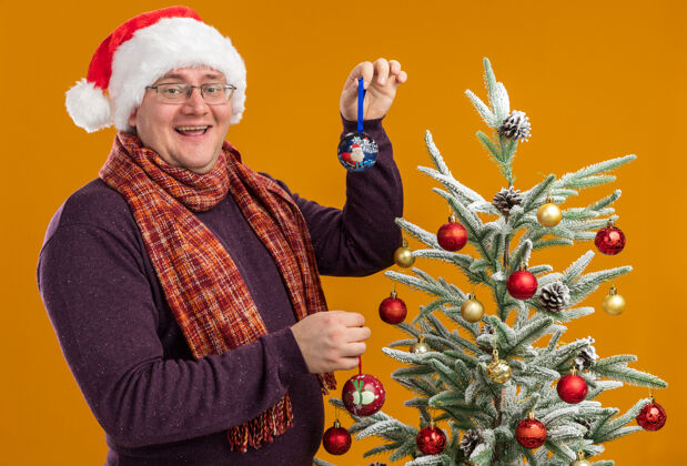 圣诞老人戴着眼镜 戴着圣诞帽 脖子上围着围巾的快乐的成年男子站在装饰过的圣诞树旁的侧视图里 手里拿着圣诞饰品 看着隔离在橙色背景上的相机附近快乐帽子