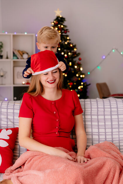坐着快乐的妈妈穿着红色的衣服坐在沙发上微笑 而她的孩子帽子母亲红色