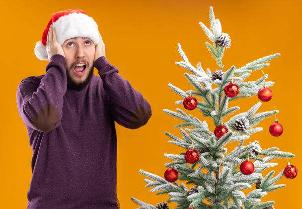 圣诞老人穿着紫色毛衣 戴着圣诞帽的年轻人站在橙色背景下的圣诞树旁 看上去既惊讶又惊讶惊讶毛衣圣诞树