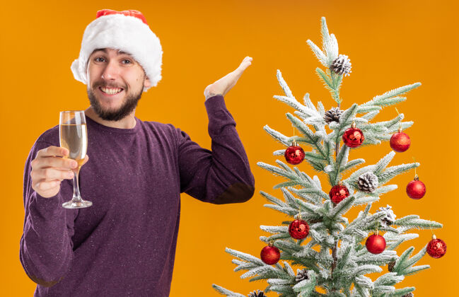年轻人一个穿着紫色毛衣 戴着圣诞帽的年轻人拿着一杯香槟看着摄像机 在橙色背景下的圣诞树旁微笑着圣诞树站着杯子