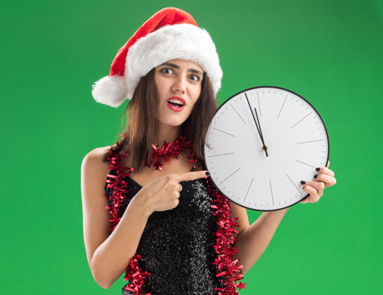 圣诞节关心的年轻漂亮女孩戴着圣诞帽 脖子上戴着花环 指着隔离在绿色墙上的挂钟花环穿关心