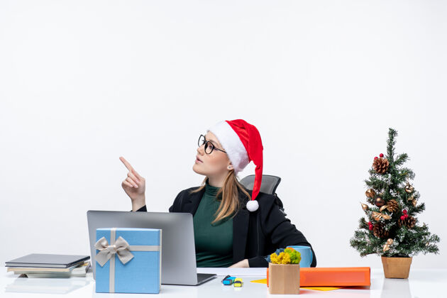 年轻一位年轻的女士拿着圣诞树和礼物坐在桌子旁 手里拿着圣诞老人的帽子和眼镜树笔记本圣诞老人