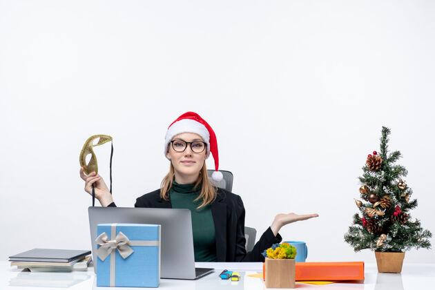 什么的戴着圣诞老人帽 戴着眼镜 拿着面具的年轻女子坐在桌子旁 手里拿着圣诞树和礼物坐着工作笔记本