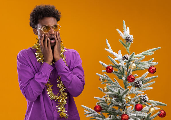 圣诞一个戴着眼镜 脖子上戴着金箔花环的美国黑人年轻人站在橙色背景的装饰过的圣诞树旁看周围金属片