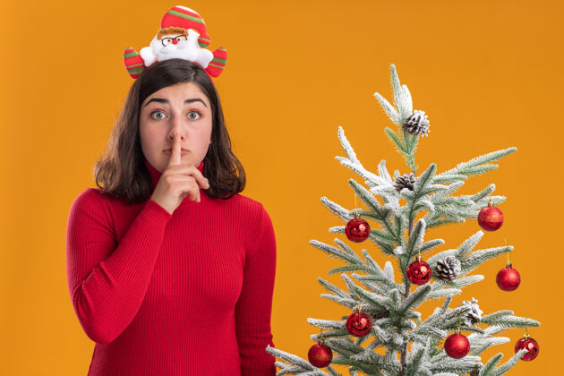 圣诞穿着圣诞毛衣的年轻女孩戴着滑稽的头带看着摄像机在橙色背景下的圣诞树旁做着沉默的手势毛衣头带穿着